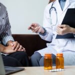 Doctor with clipboard discussing with patient, medication bottles on table, laptop in foreground.